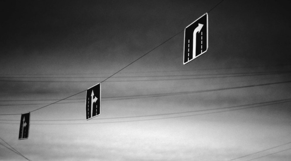 Drei Richtungsschilder in schwarz-weiss an einer Oberleitung aufgehängt